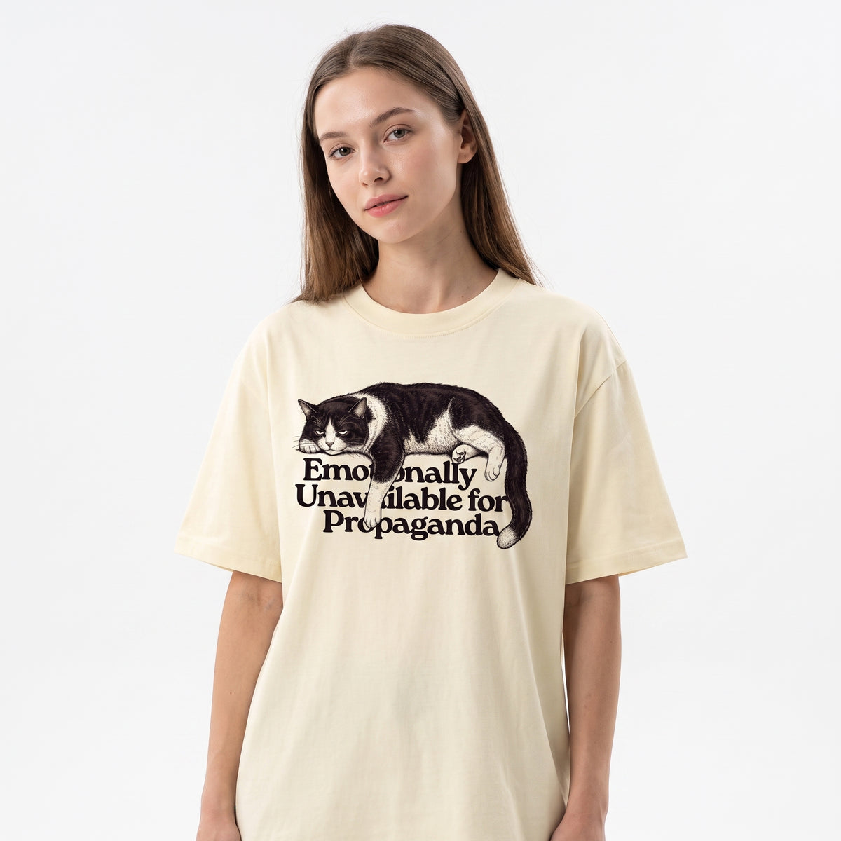 A woman wearing a cream-colored oversized t-shirt with a graphic of a sleeping black and white cat and the text 'Emotionally Unavailable for Propaganda'.