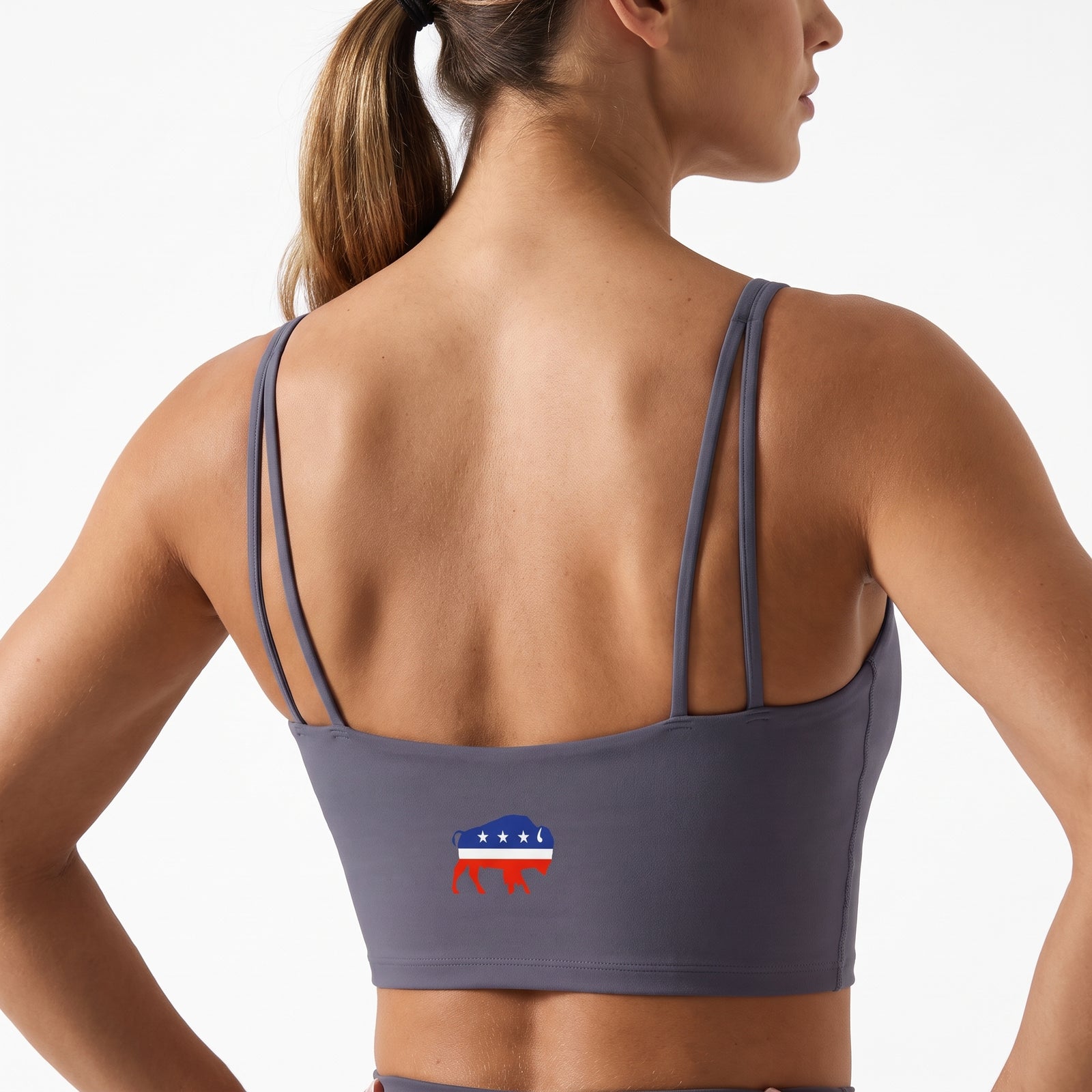 Rear view of a woman wearing a dark gray sports bra with a small red, white, and blue graphic on the back, set against a plain white background.