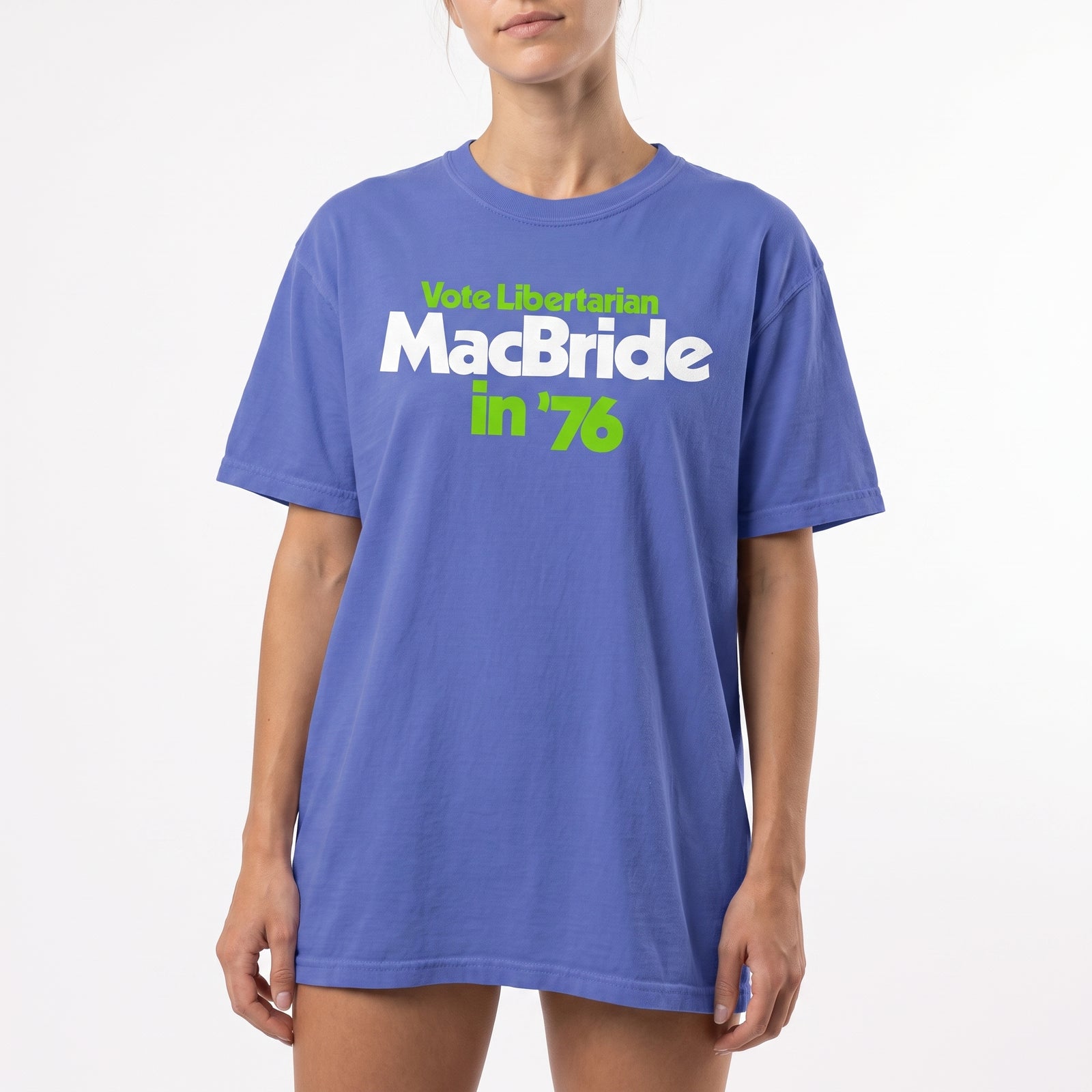 A woman wearing an oversized blue t-shirt with a political graphic that reads 'Vote Libertarian MacBride in '76' in white and lime green text, set against a plain white background.
