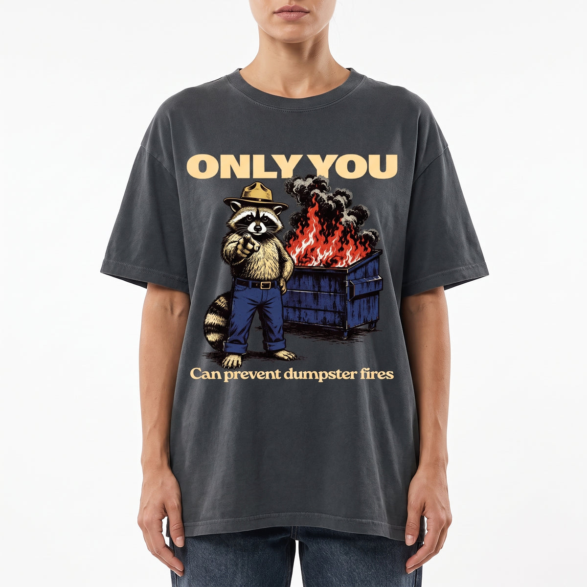 A model wearing a dark gray oversized graphic t-shirt. The shirt features a cartoon raccoon wearing a fire department hat standing next to a dumpster on fire, with the text 'ONLY YOU' above and 'Can prevent dumpster fires' below.