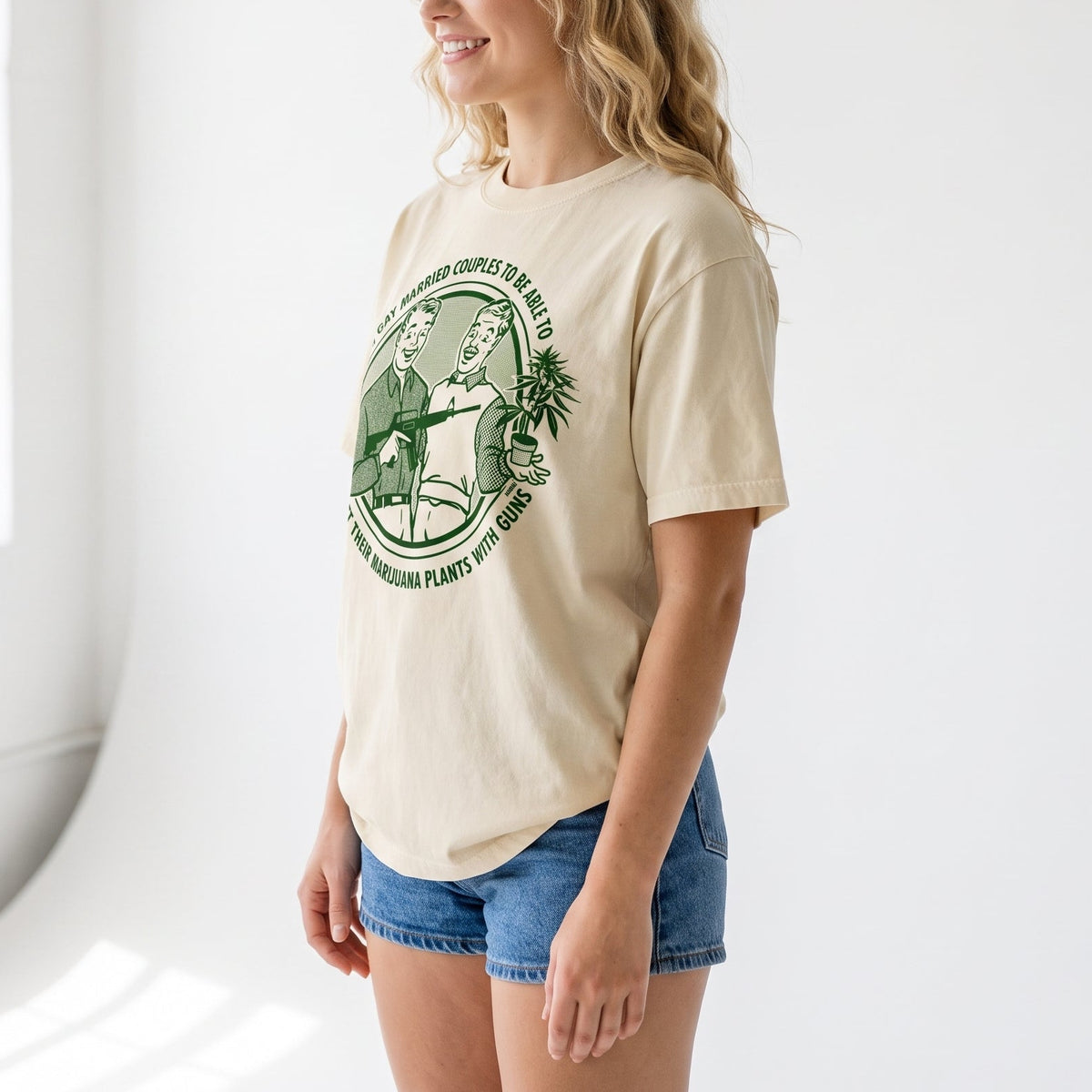 A woman with blonde wavy hair stands in a bright white studio setting, wearing a cream-colored t-shirt with a green circular graphic print and blue denim shorts.