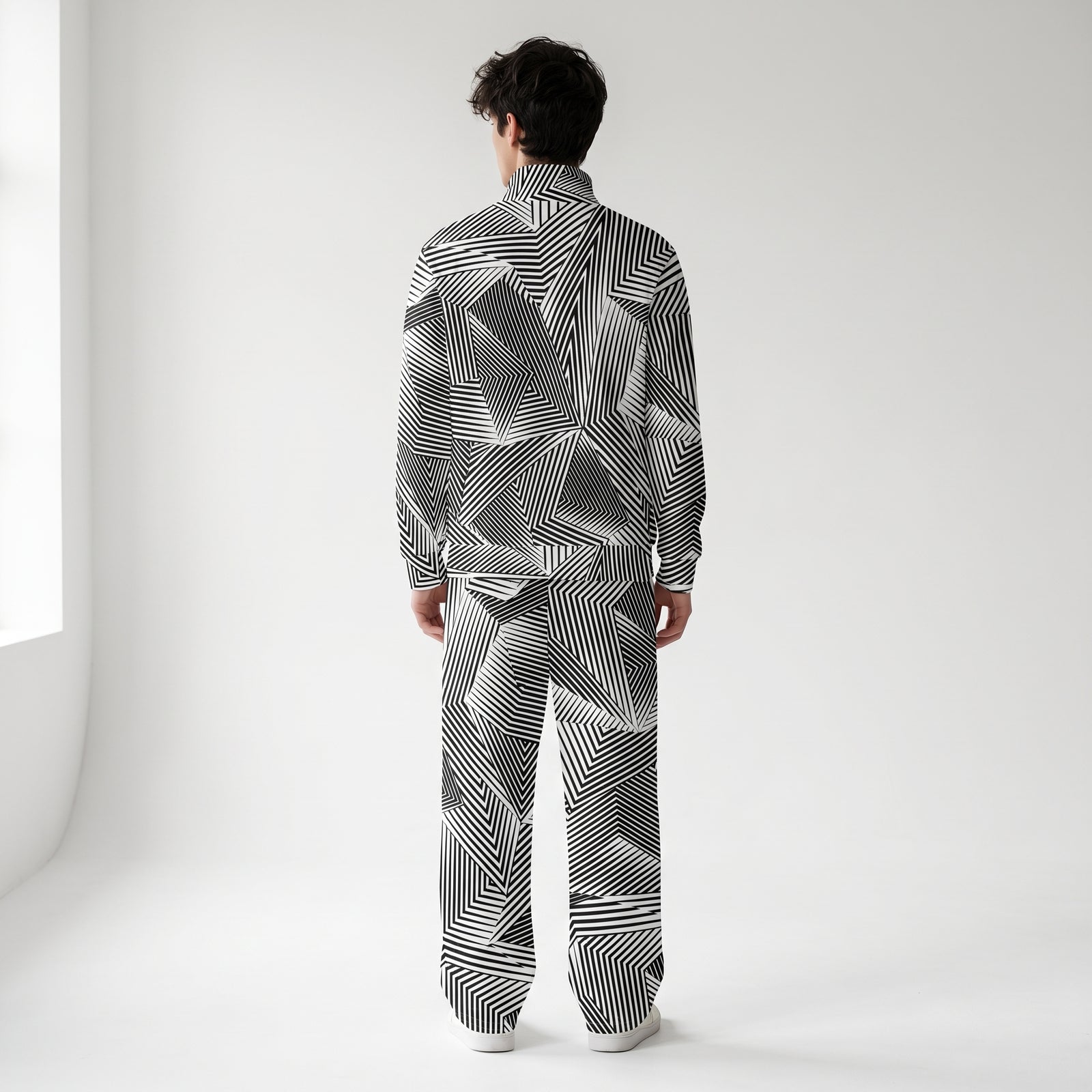 A man viewed from behind wearing a matching Liberty Maniacs Dazzle Naval Camouflage long-sleeve track jacket and wide-leg pants set with a bold black and white dazzle geometric stripe pattern, standing in a bright white studio. 