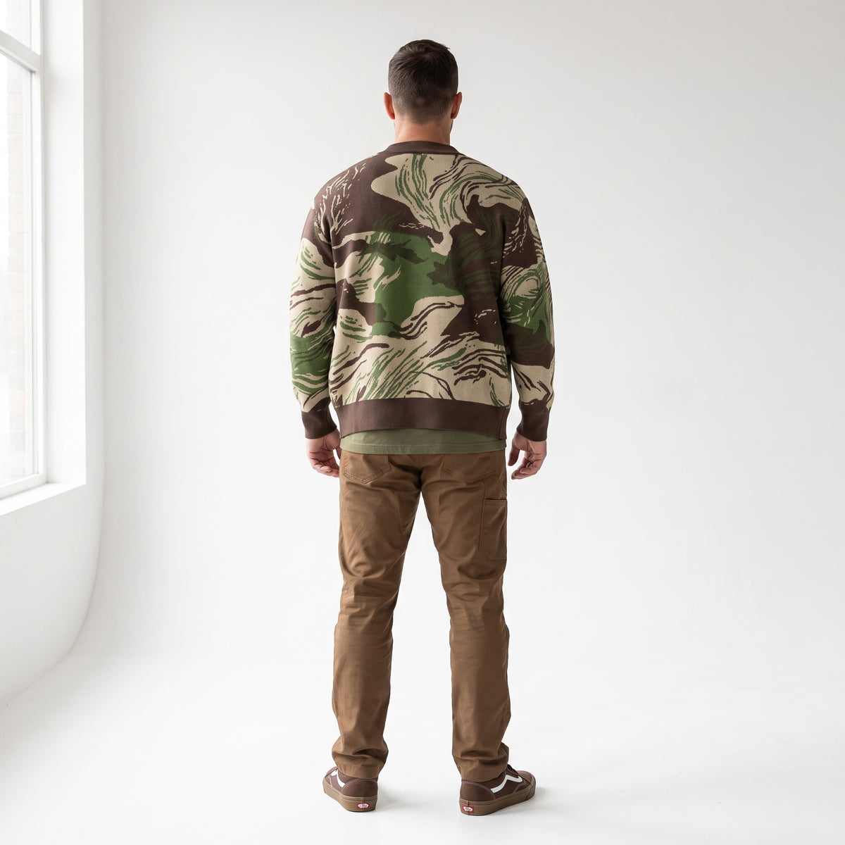 A man viewed from behind wearing a Rhodesian Brushstroke Knitted Cardigan with a green and brown abstract camouflage pattern, paired with brown pants and brown shoes, standing in a bright white room.