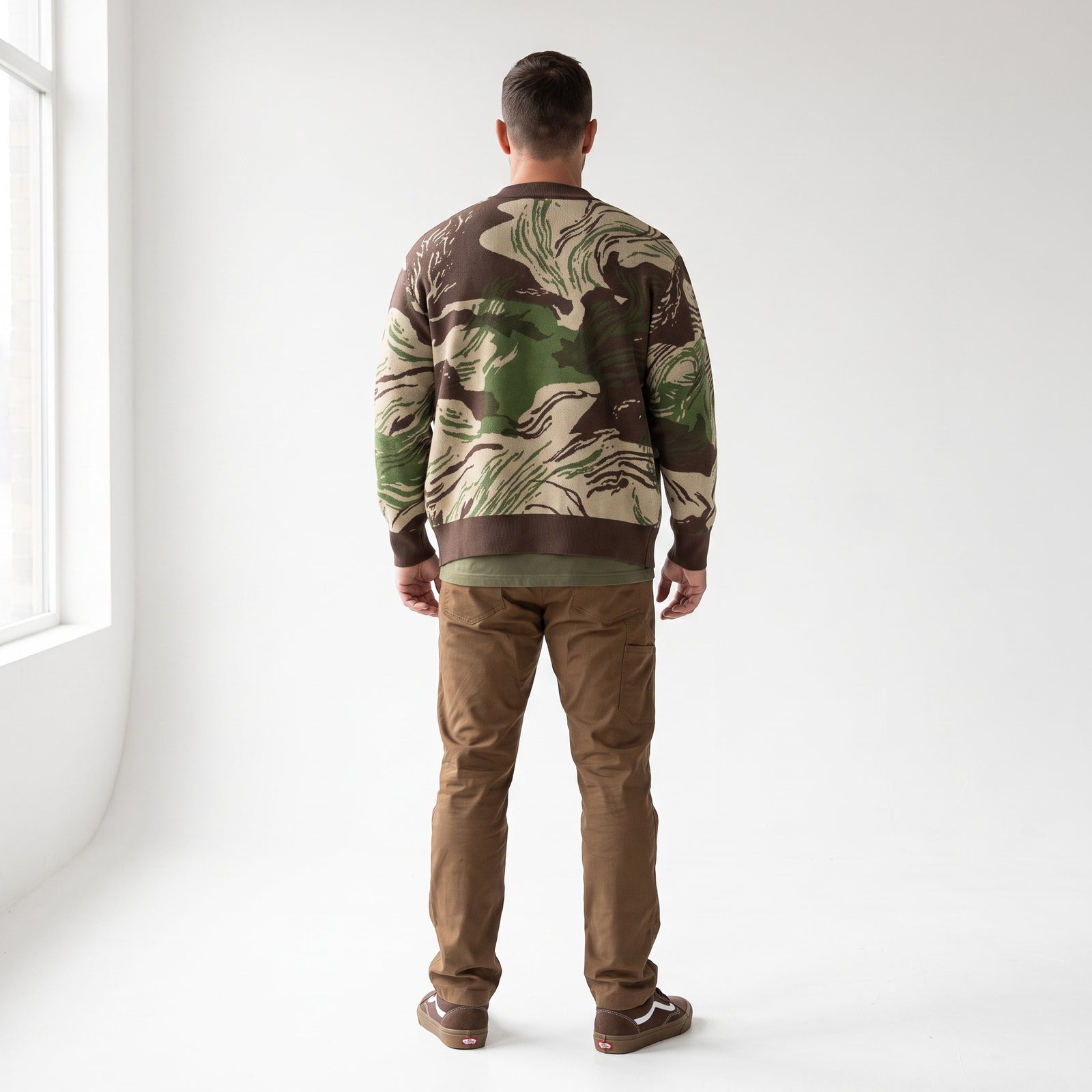 A man viewed from behind wearing a Rhodesian Brushstroke Knitted Cardigan with a green and brown abstract camouflage pattern, paired with brown pants and brown shoes, standing in a bright white room.