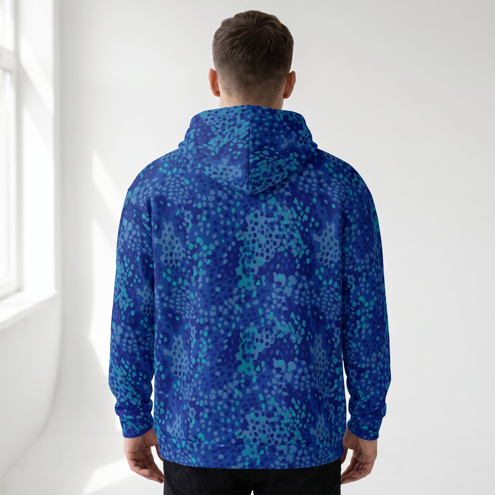 A man viewed from behind wearing a blue and teal patterned hoodie with a dotted abstract design, set against a bright white wall with soft window light.