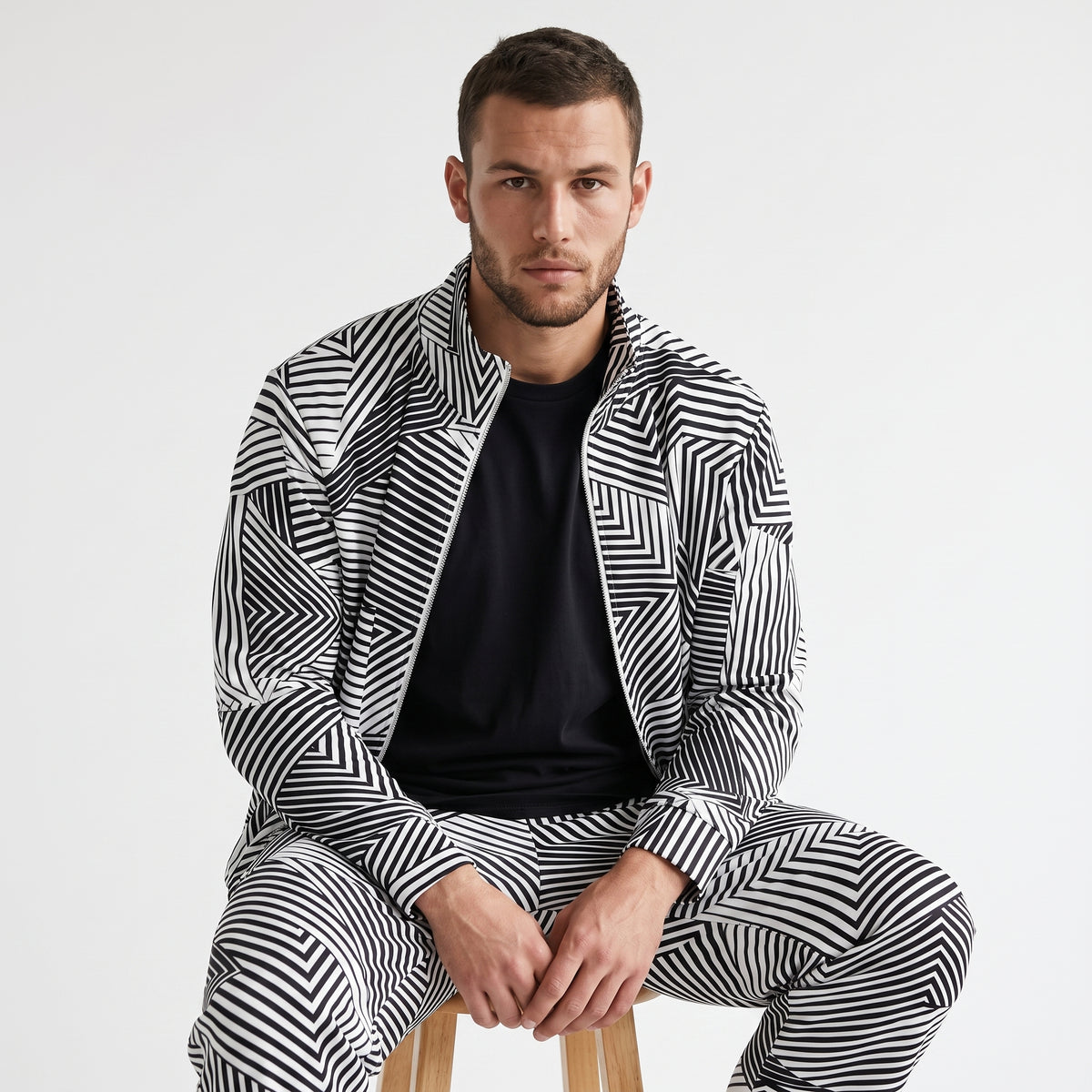 A man wearing a matching black and white geometric patterned zip-up jacket and trousers set, layered over a black t-shirt, sitting on a wooden stool against a plain white studio background.