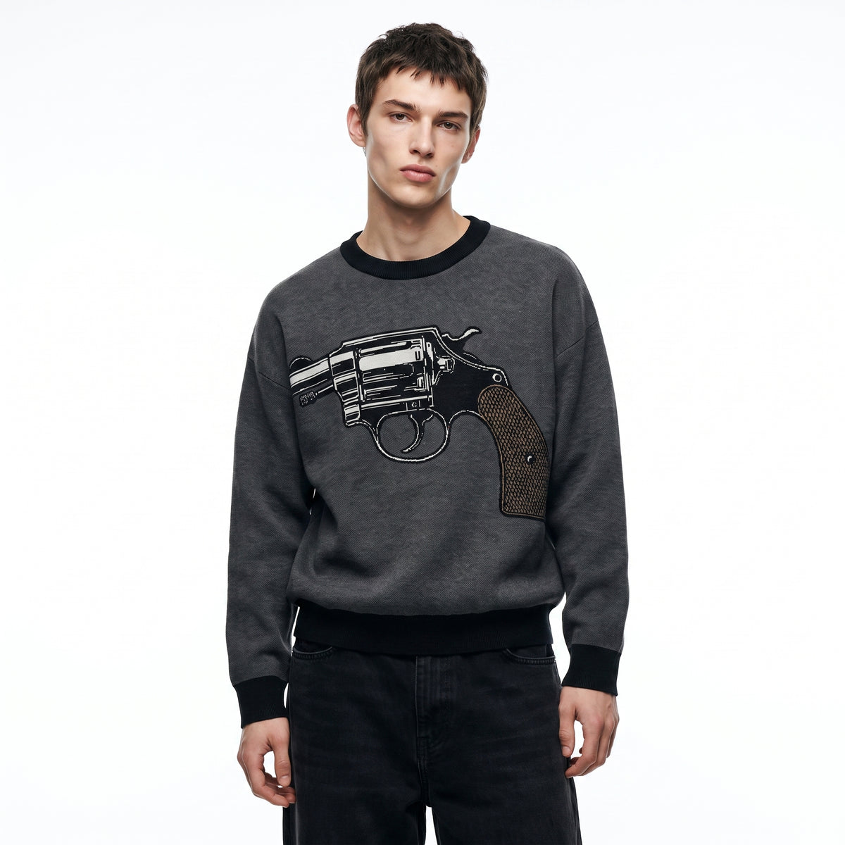 A man wearing a dark gray crewneck knit sweater with a large black and silver revolver graphic on the front, paired with black jeans, against a plain white background.