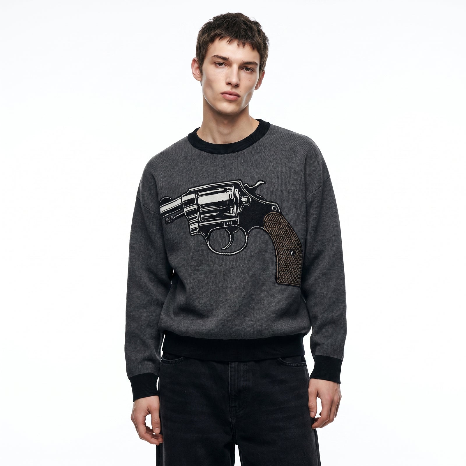 A man wearing a dark gray crewneck knit sweater with a large black and silver revolver graphic on the front, paired with black jeans, against a plain white background.