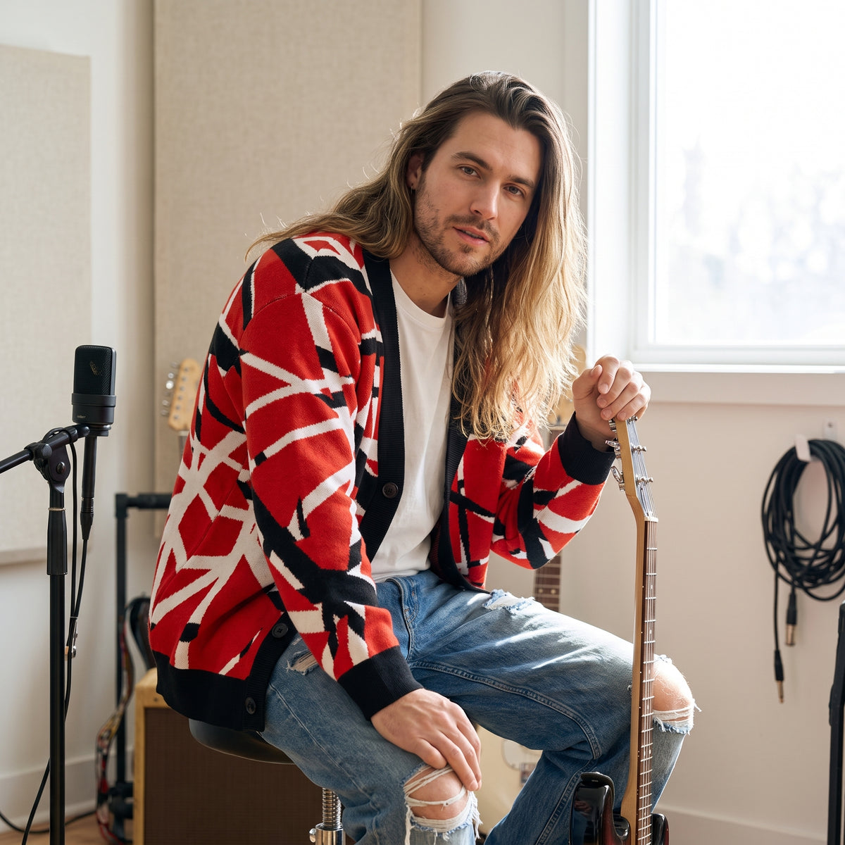 A man with long blonde hair sits in a music studio wearing a red, black, and white geometric patterned cardigan over a white t-shirt and ripped blue jeans. He is holding an electric guitar, with microphones and studio equipment visible in the background.