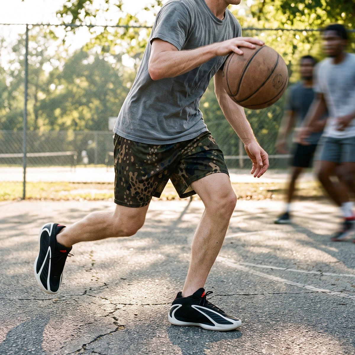 A man dribbling a basketball on an outdoor court at sunset, wearing a gray t-shirt, camouflage-patterned shorts, and black and white sneakers. Two other players are blurred in the background near a chain-link fence and trees.