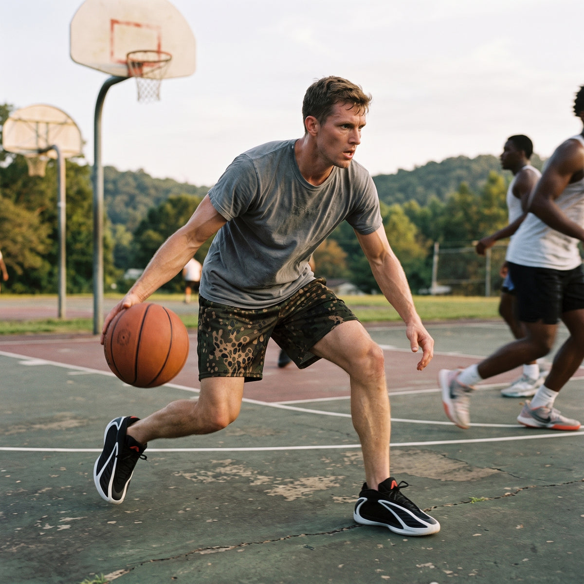 A man dribbling a basketball on an outdoor basketball court, wearing a gray t-shirt, Erbsenmuster Pea Dot Field Green Camo Mesh Shorts, and black and white sneakers. Other players are visible in the background, along with trees and a basketball hoop.