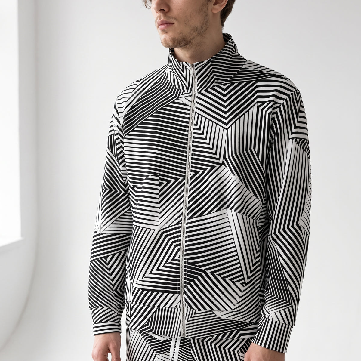 A man wearing a black and white Dazzle Camouflage geometric patterned zip-up track jacket and matching pants, shown from the side against a bright white background.