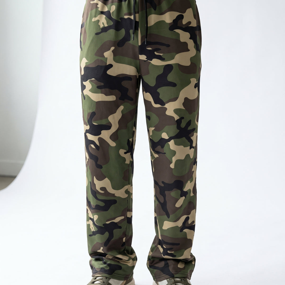 A pair of camouflage sweatpants shown from the waist down, featuring a classic green, brown, black, and beige camo pattern. The pants have a drawstring waist and are paired with light-colored sneakers, photographed against a clean white studio background.