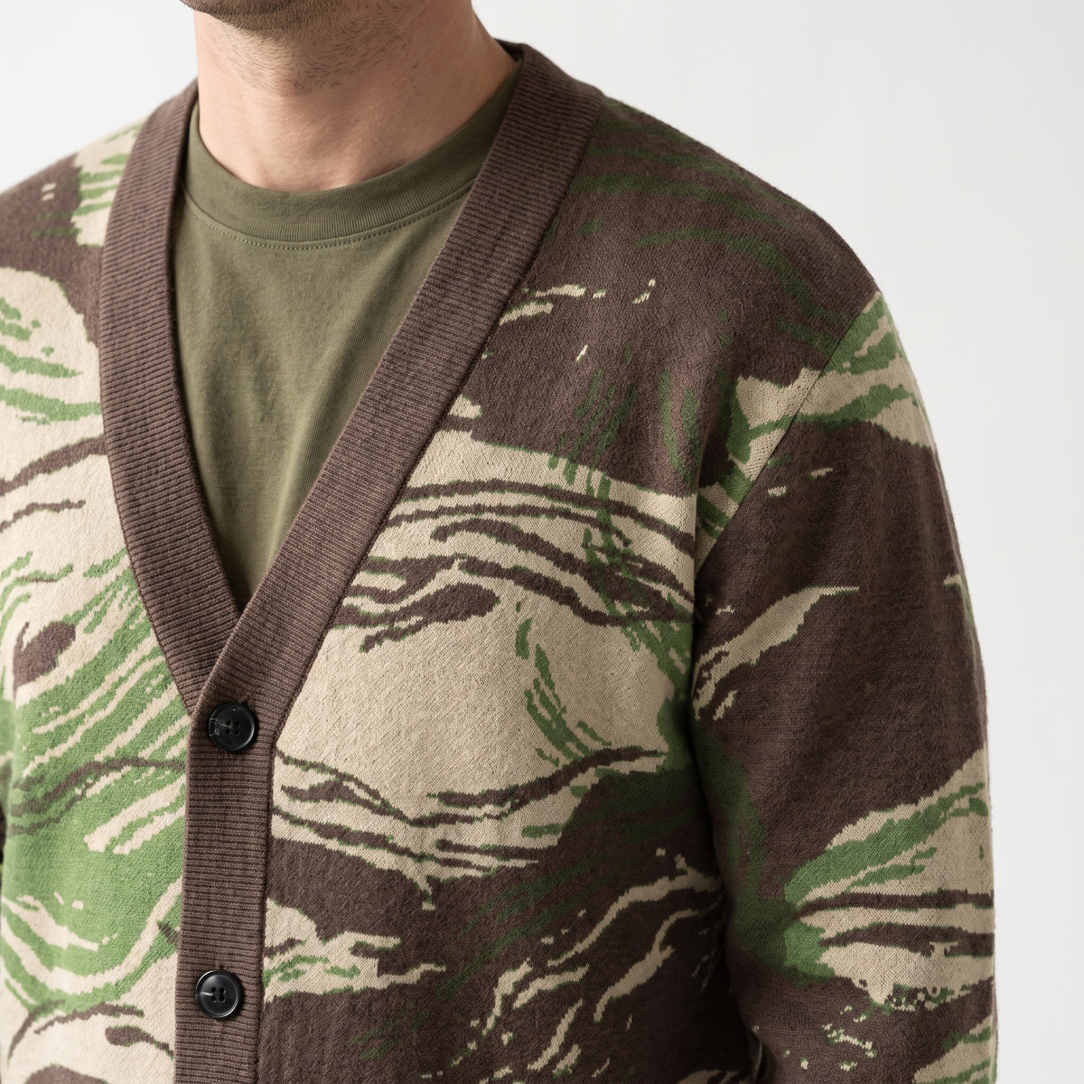 A close-up of a man wearing a brown, beige, and greenRhodesian Brushstroke Knitted Cardigan over an olive green t-shirt, against a plain light background.