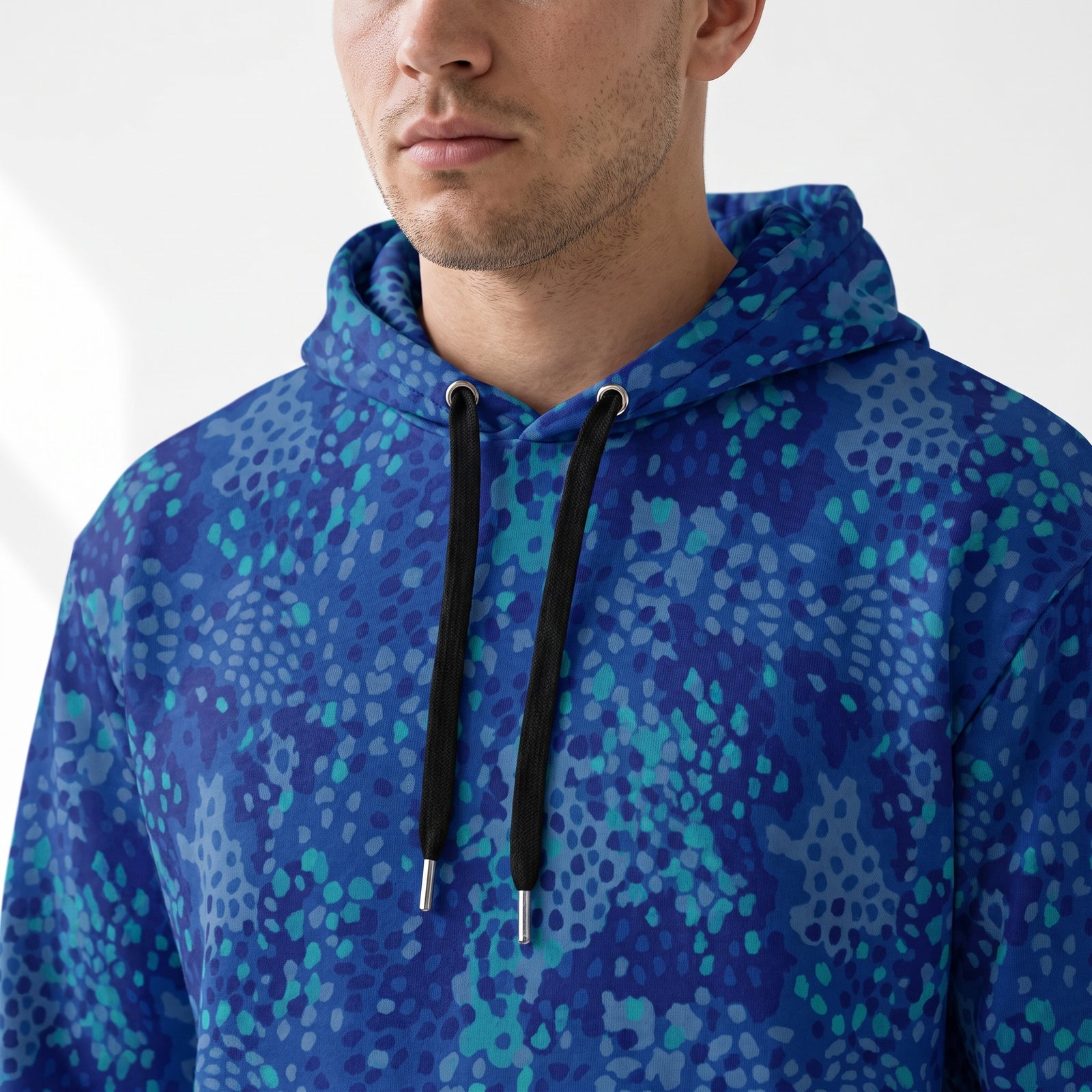 A close-up of a man wearing a blue and teal abstract patterned hoodie with black drawstrings, shown from the chest up against a clean white background.