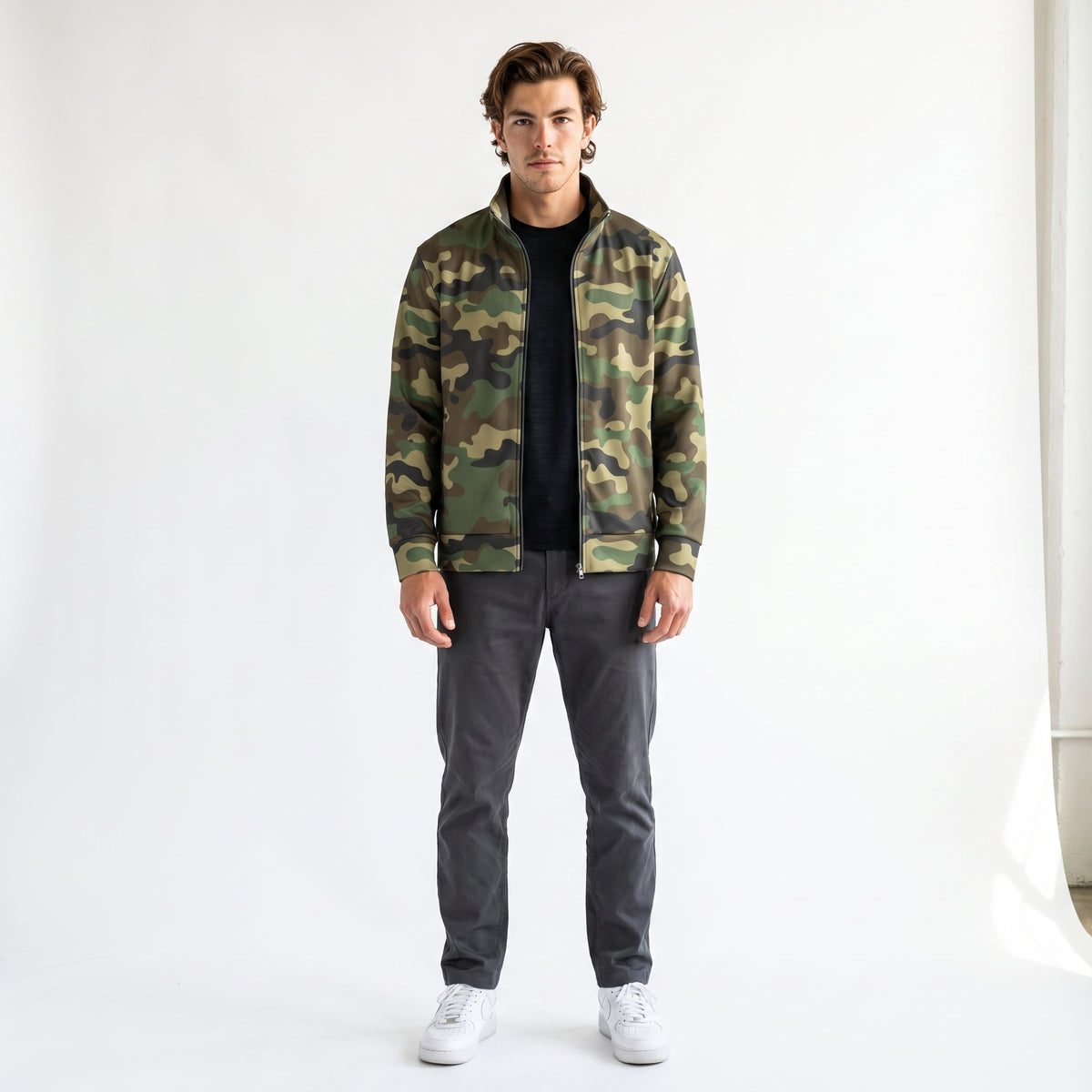 A man modeling a green and brown camouflage bomber jacket over a black t-shirt, dark gray pants, and white sneakers, standing against a plain white studio background.