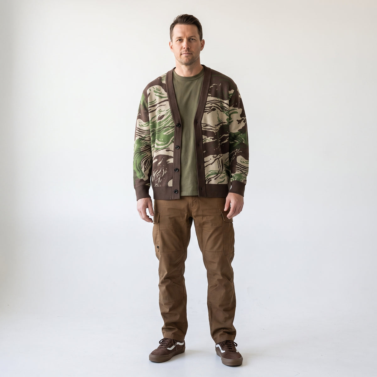 A man modeling a brown and green Rhodesian Brushstroke Knitted Cardigan over an olive green t-shirt and brown pants, standing against a plain white studio background.