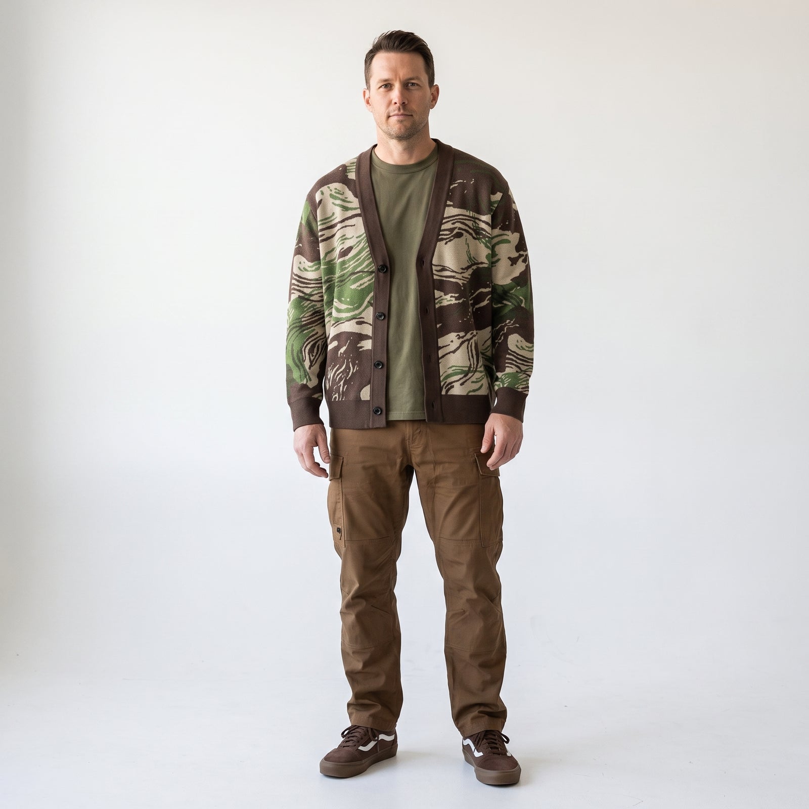 A man modeling a brown and green Rhodesian Brushstroke Knitted Cardigan over an olive green t-shirt and brown pants, standing against a plain white studio background.