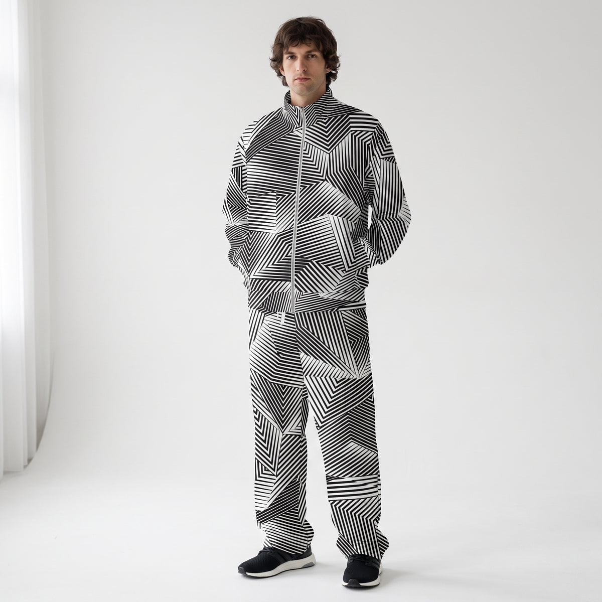 A man wearing a matching zip-up jacket and pants set with a black and white geometric striped pattern, standing against a plain light gray studio background.