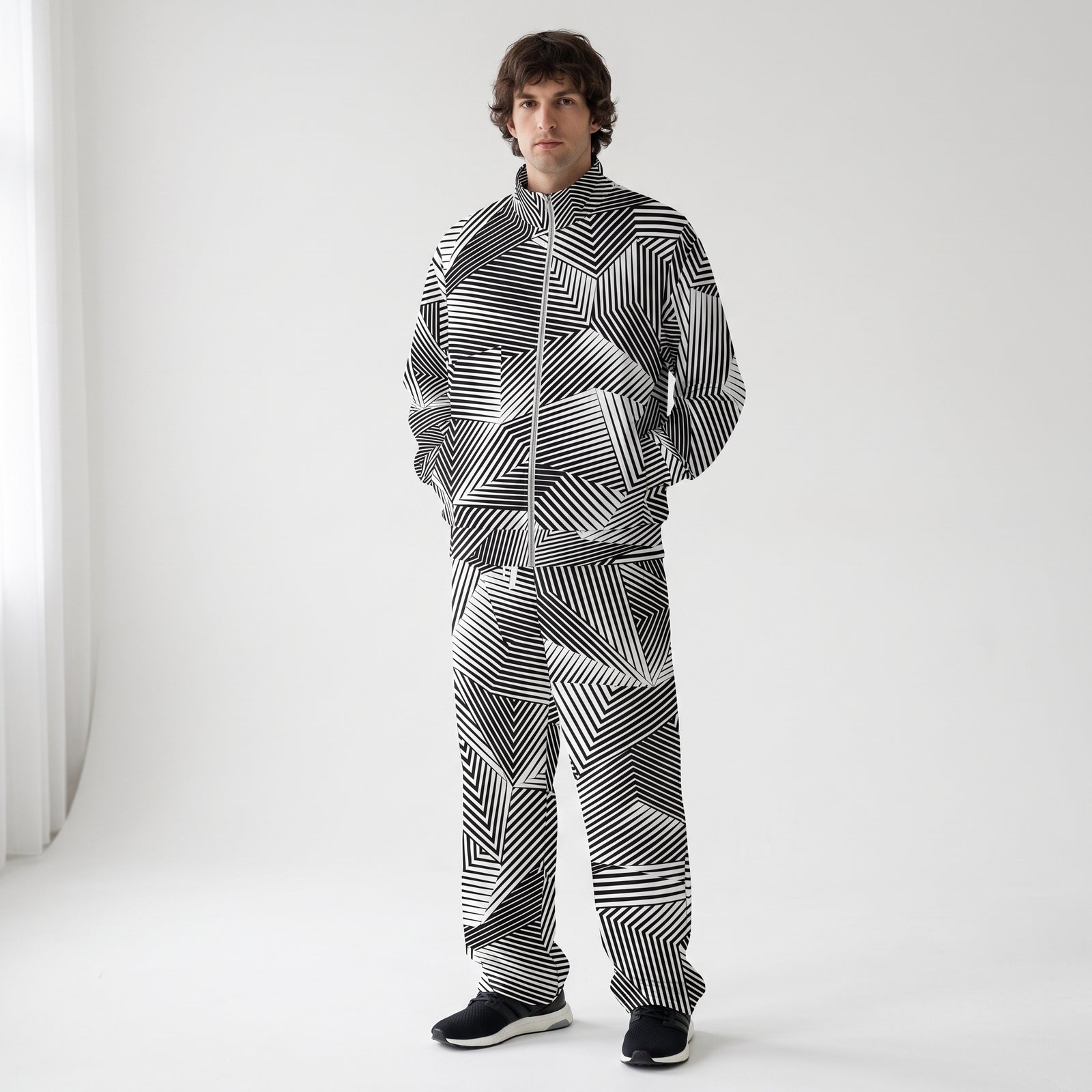 A man wearing a matching zip-up jacket and pants set with a black and white dazzle geometric striped pattern, standing against a plain light gray studio background.