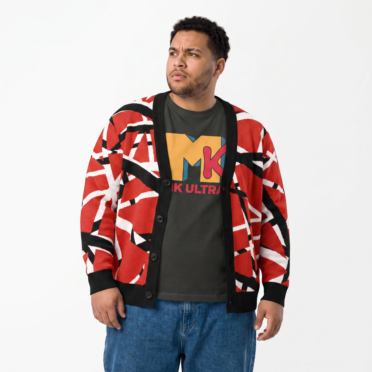A man wearing a red, black, and white geometric patterned cardigan over a dark olive graphic t-shirt and blue jeans, standing against a plain white studio background.