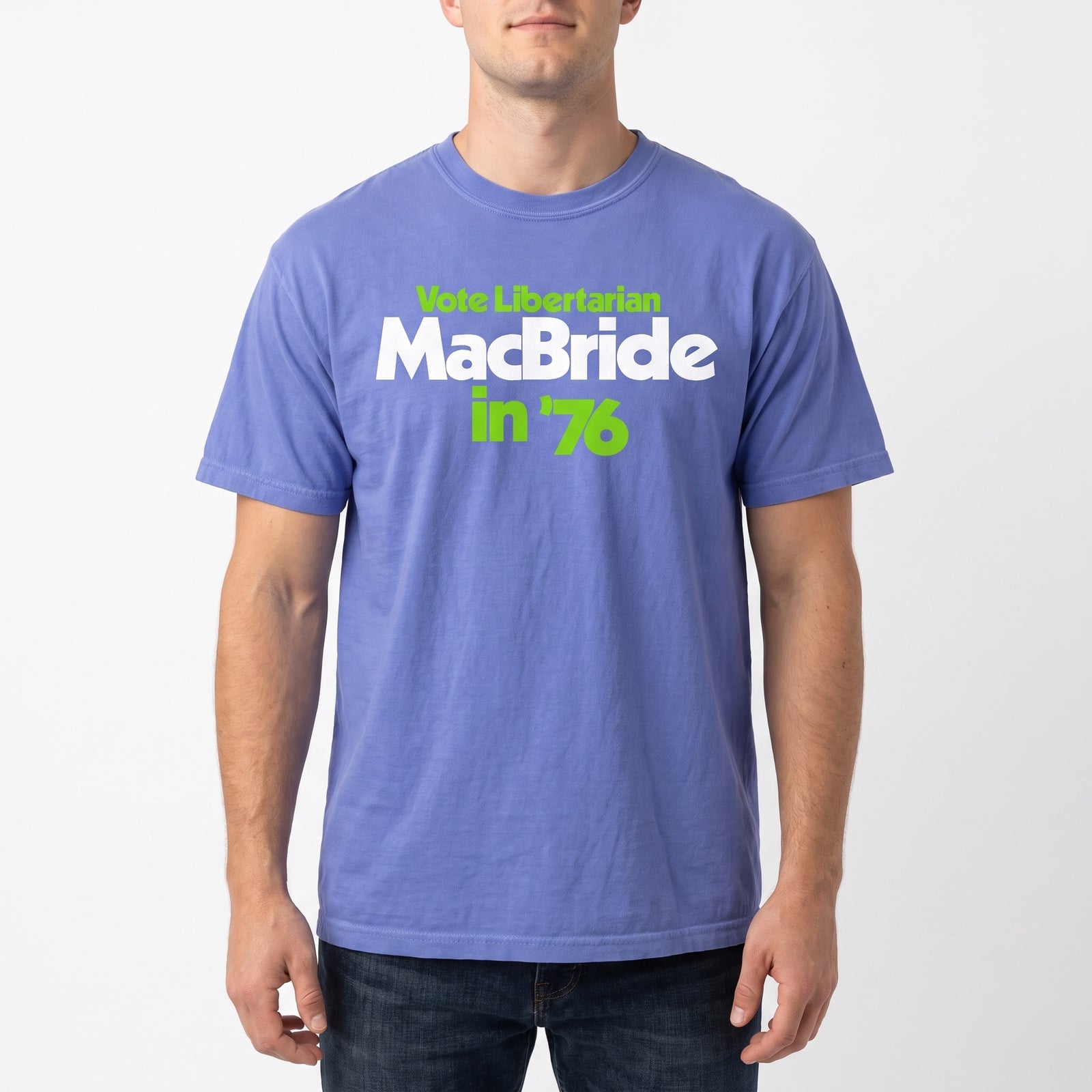 A man wearing a heathered periwinkle blue t-shirt with a graphic print that reads 'Vote Libertarian MacBride in '76' in white and lime green text, paired with dark blue jeans against a light gray background.