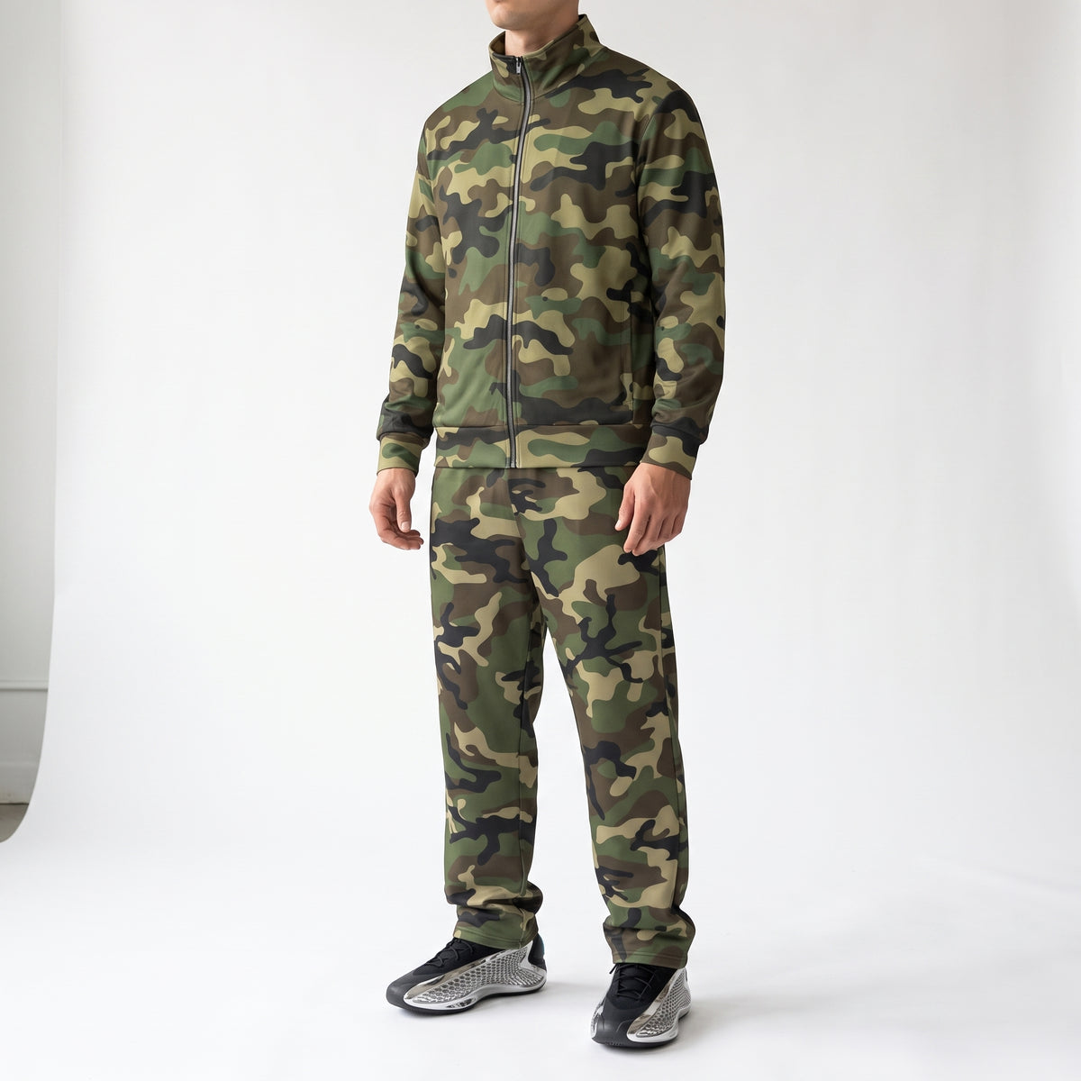 A model wearing a matching camouflage tracksuit, featuring a zip-up jacket and jogger pants, paired with black and gray sneakers, photographed against a white studio background.