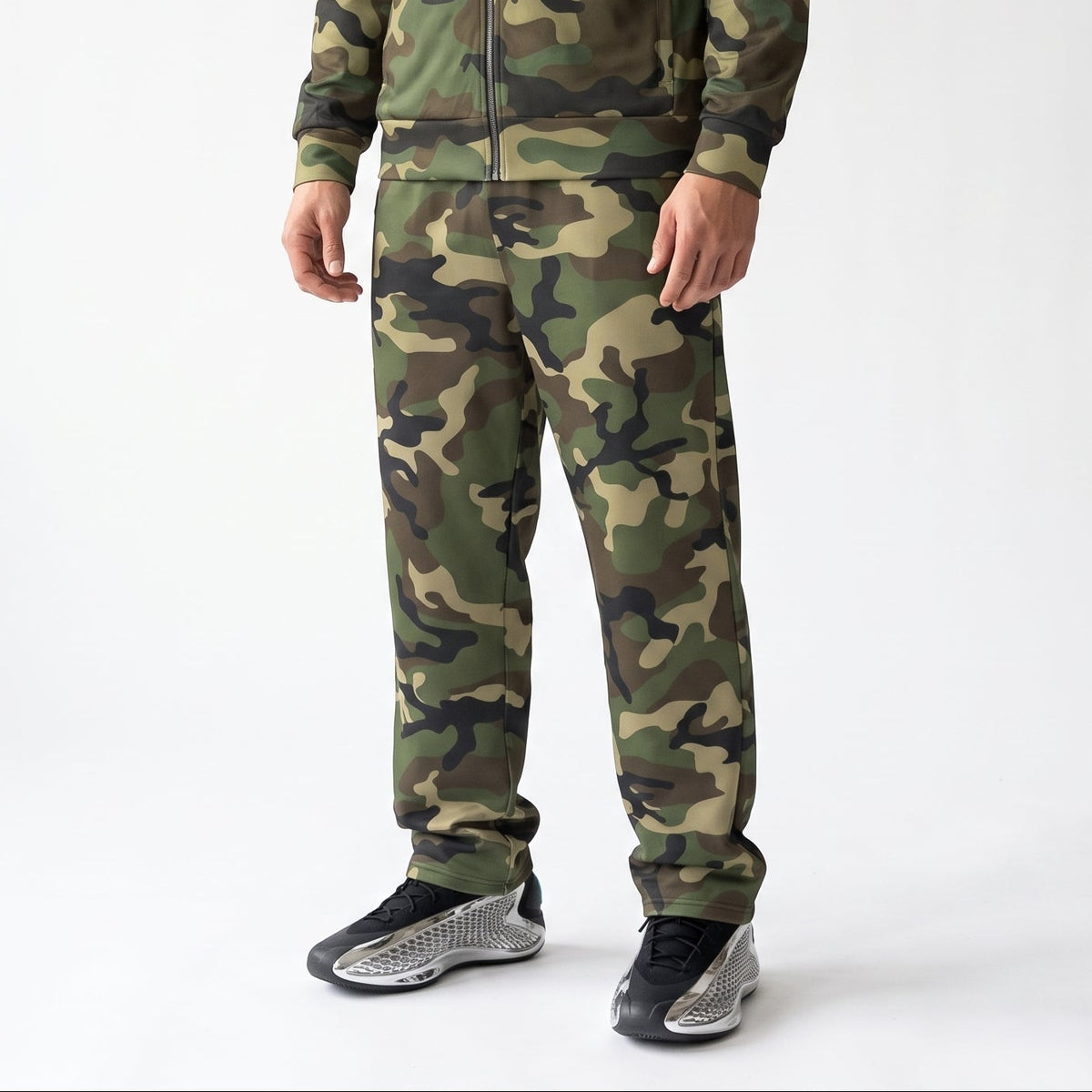 A model wearing a matching camouflage tracksuit, featuring a zip-up jacket and jogger pants, paired with black and gray sneakers, photographed against a white studio background.