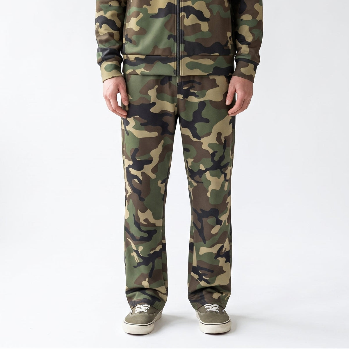 A man wearing a matching camouflage tracksuit, consisting of a zip-up jacket and matching pants, paired with gray sneakers, standing against a plain white studio background.