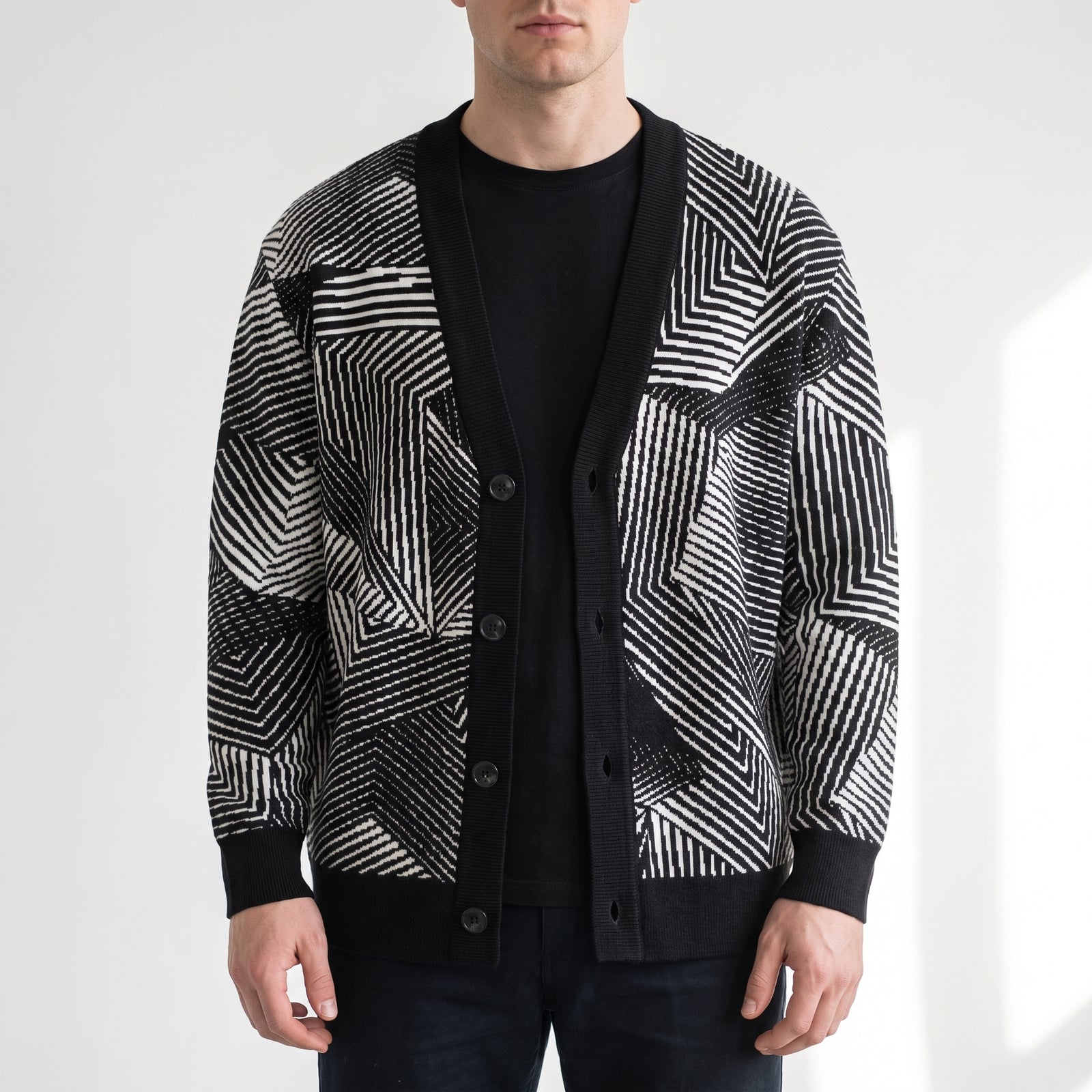 A man wearing a black and white geometric patterned knit cardigan over a black crewneck t-shirt and dark jeans, photographed against a clean white studio background.