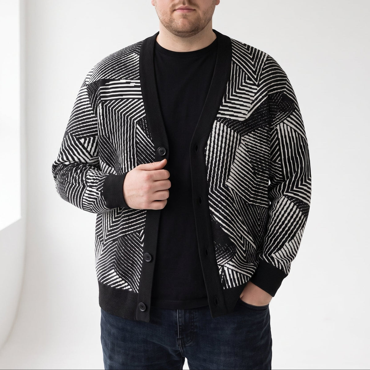 A man wearing a black and white geometric patterned cardigan over a black t-shirt and dark jeans, standing in a bright white room.