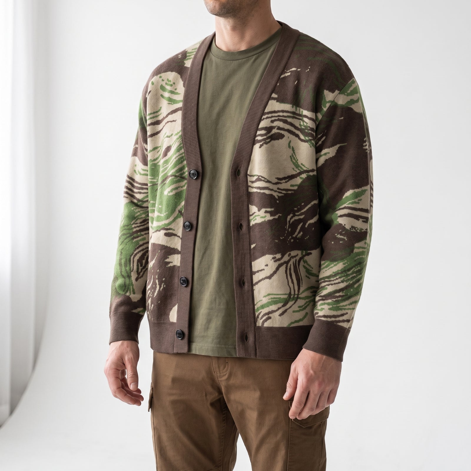 A man wearing a brown and green camouflage knit cardigan over an olive green t-shirt and brown pants, photographed in a bright studio setting.