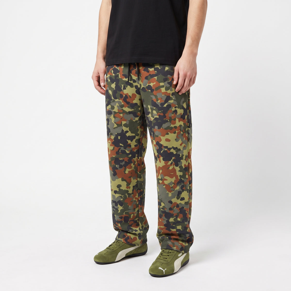 A person wearing camouflage jogger pants with a black t-shirt and olive green Puma sneakers, shown from the waist down on a light gray studio background.