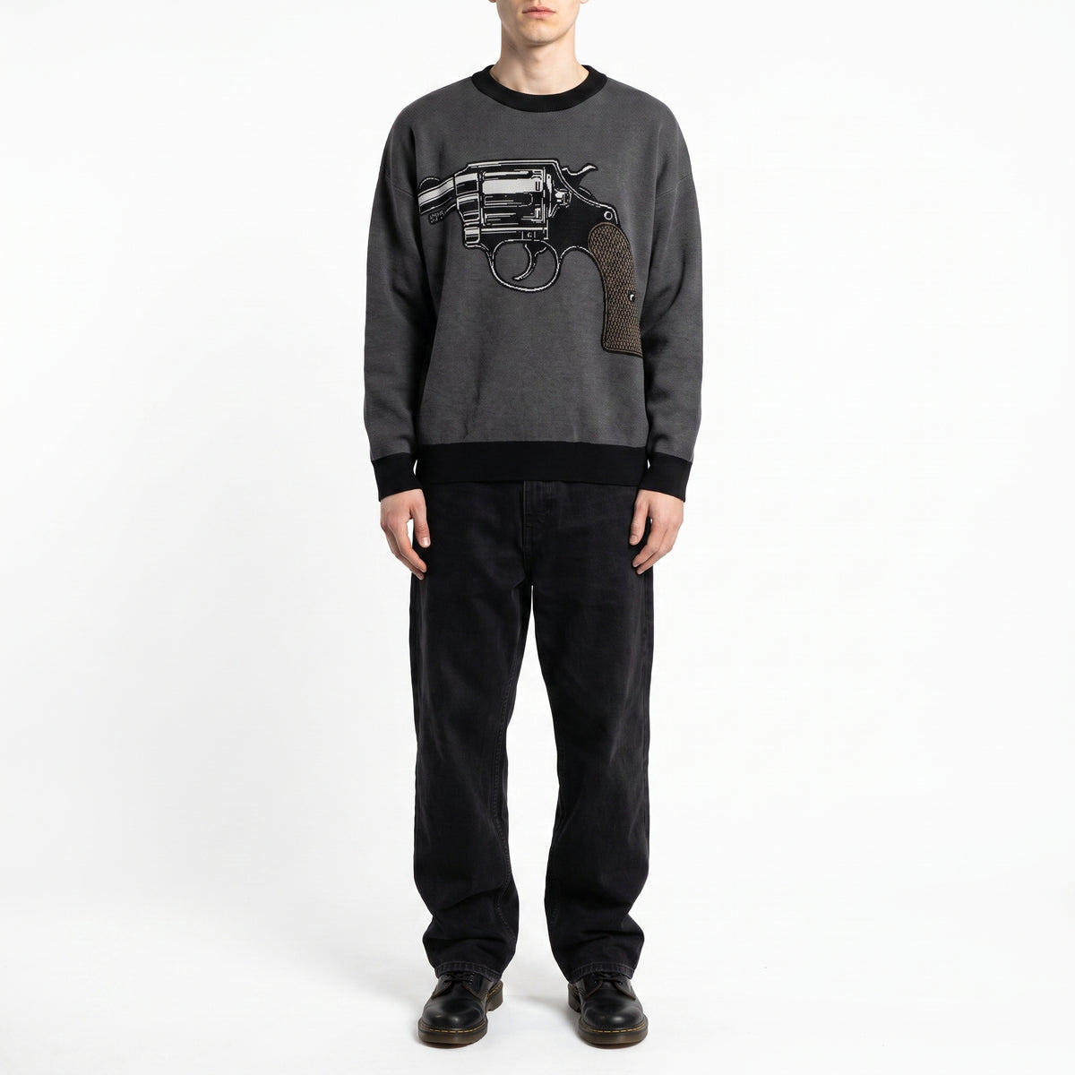 A model wearing a dark gray crewneck Jacquard-knit sweater with a large black and brown revolver graphic on the chest, paired with black pants and black shoes, shot against a plain white studio background.