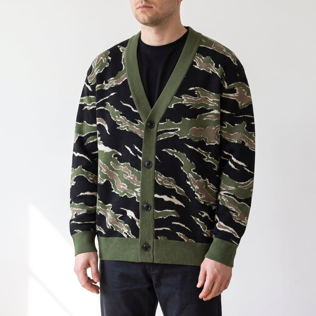 A man wearing a green and black tiger-stripe patterned cardigan over a black t-shirt and dark jeans, photographed against a clean white studio background.