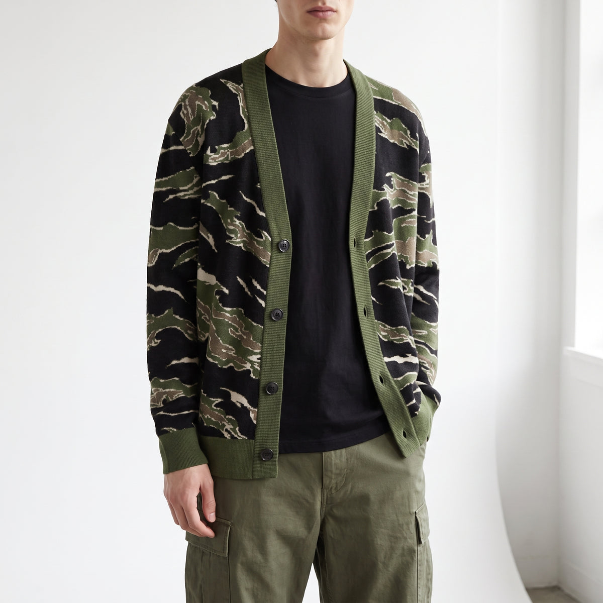 A man wearing a green and black tiger-stripe patterned cardigan over a black t-shirt and olive green cargo pants, standing in a brightly lit room.