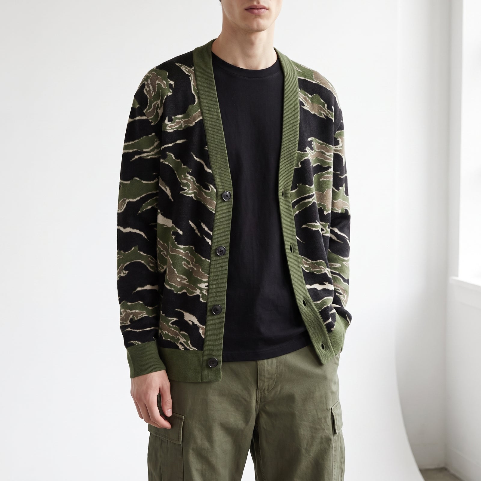A man wearing a green and black tiger-stripe patterned cardigan over a black t-shirt and olive green cargo pants, standing in a brightly lit room.