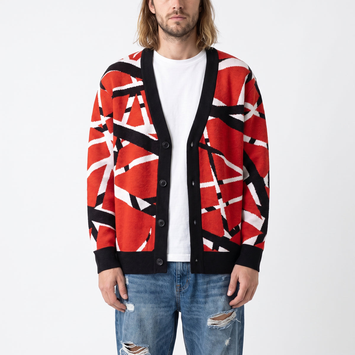 A man wearing a red open-front cardigan with a black and white abstract geometric pattern, styled over a white t-shirt and distressed blue jeans against a plain white background.