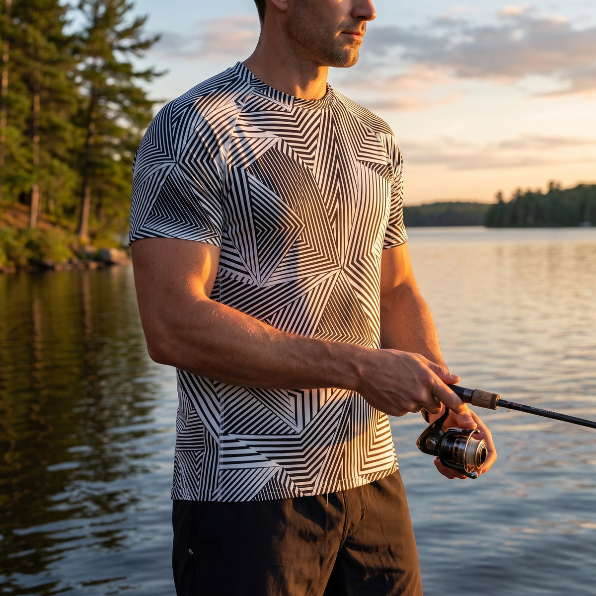 A man fishing by a lake at sunset, wearing a short-sleeve athletic t-shirt with a black, white, and gray geometric pattern.
