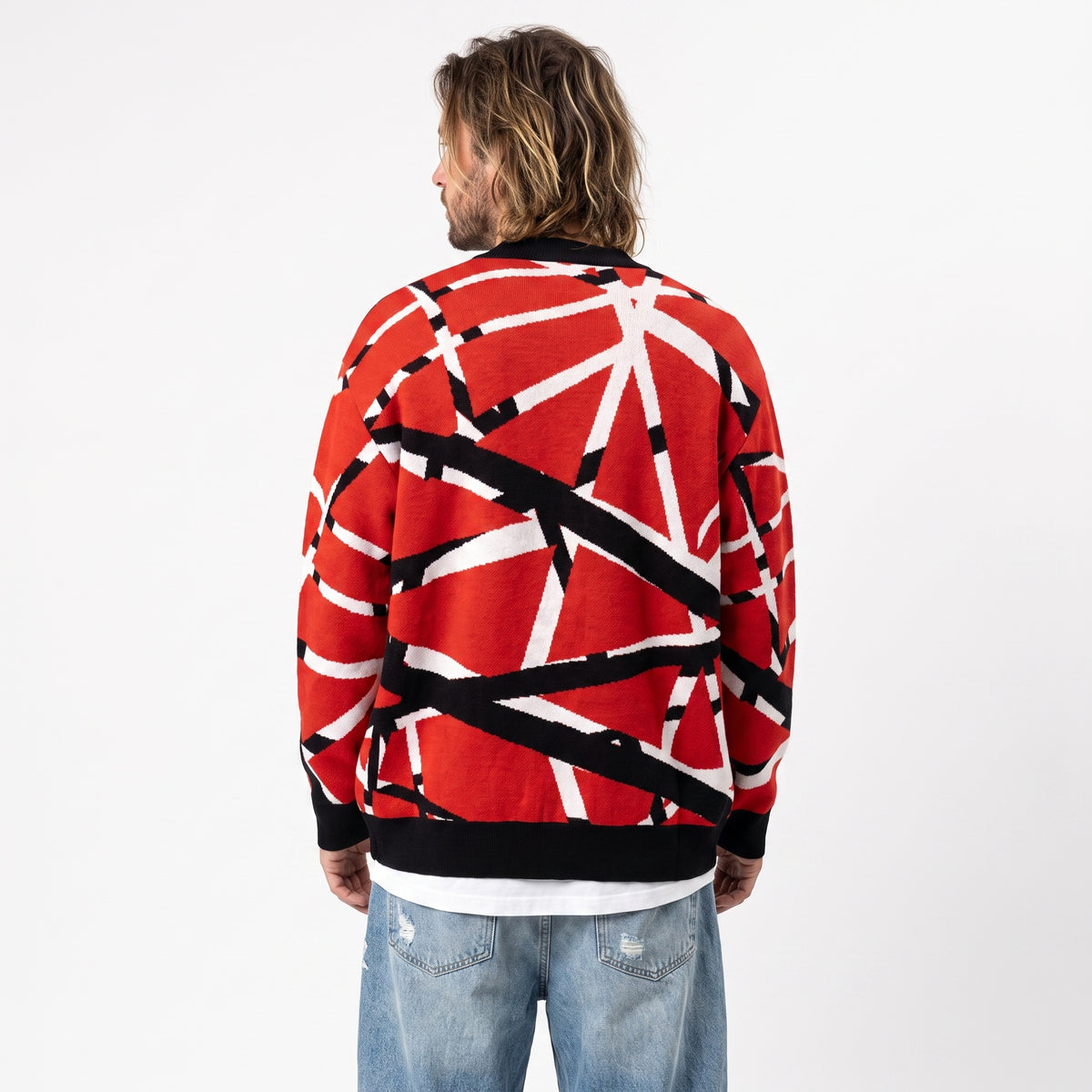 A man viewed from behind wearing a red knit sweater with a bold black and white abstract pattern, paired with light blue jeans, against a plain white studio background.