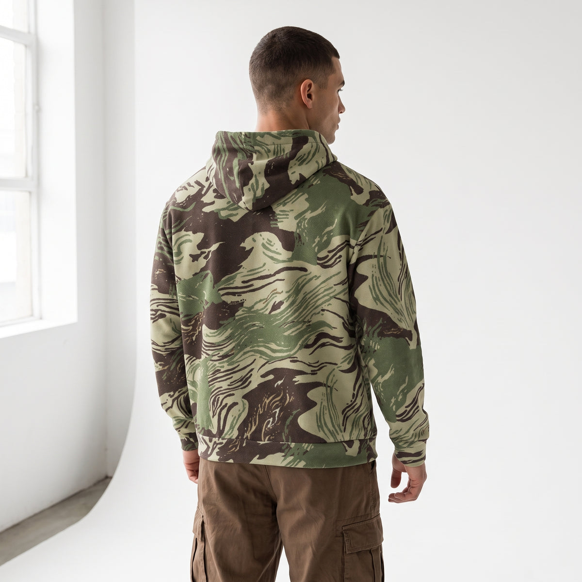 A man viewed from behind wearing a green and brown camouflage hoodie and brown cargo pants, standing in a bright white room.