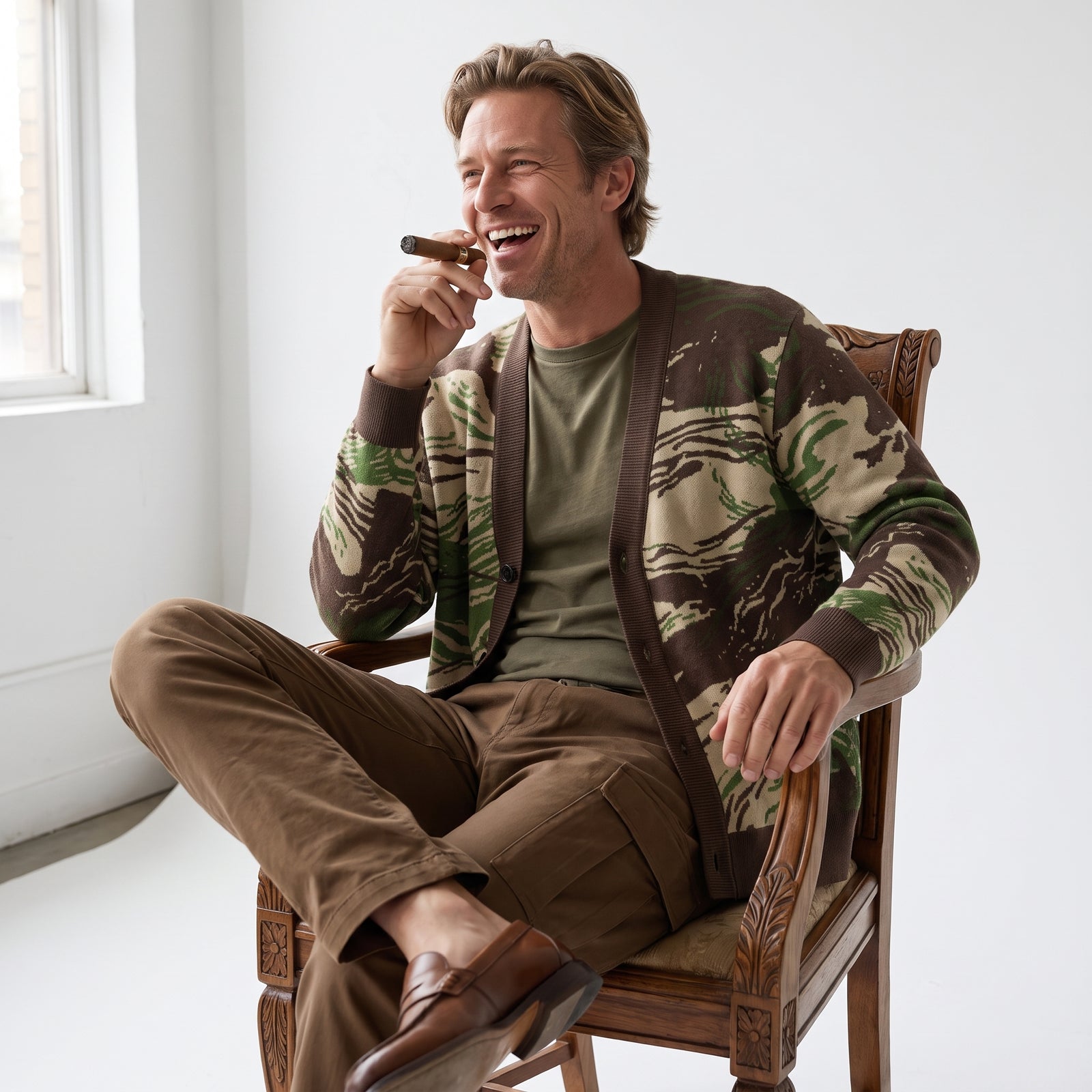 A man sitting in a wooden chair, wearing a brown and green Rhodesian Brushstroke Knitted Cardigan over an olive green t-shirt and brown trousers. He is holding a cigar near his face in a bright, minimalist room.