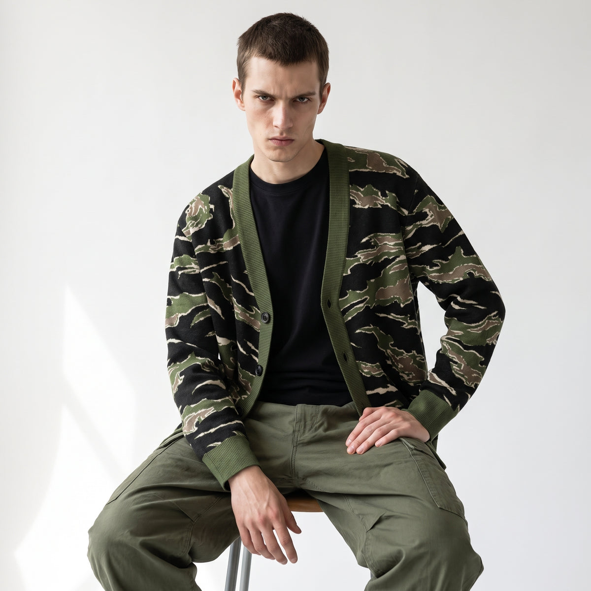 A man wearing a green and black tiger stripe camouflage cardigan over a black t-shirt and olive green cargo pants, sitting on a stool against a white studio background.