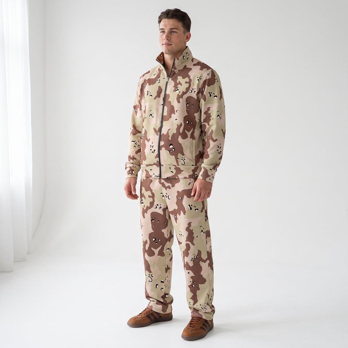 A man modeling a matching two-piece tracksuit in a desert camouflage pattern, standing against a bright white studio background.