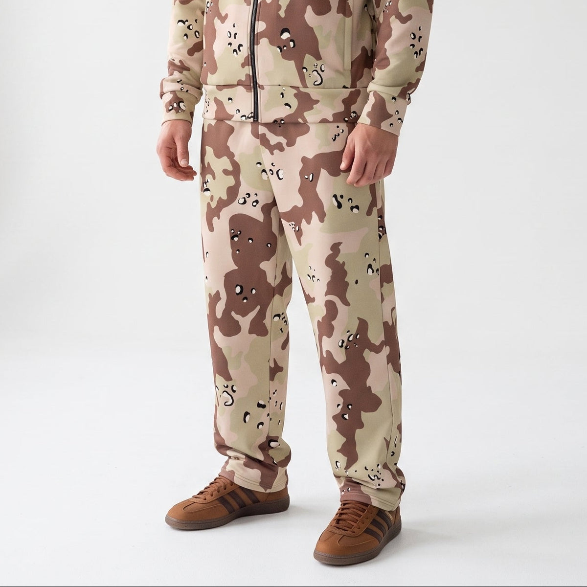 A man modeling a matching two-piece tracksuit in a desert camouflage pattern, standing against a bright white studio background.
