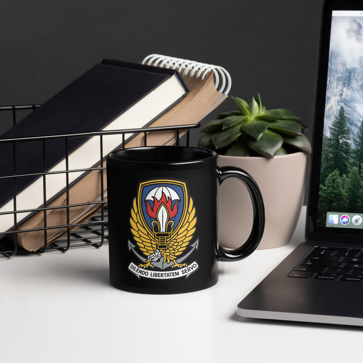 Black mug with a colorful emblem on a desk with a laptop and books.