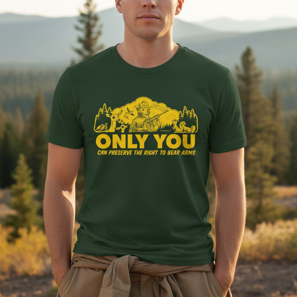 A dark green t-shirt from Liberty Maniacs, folded to show the graphic. The design features a yellow cartoon bear, a parody of Smokey Bear, holding an assault rifle. He is in a forest with a deer, squirrels, and birds. Below the image, the text reads, &quot;ONLY YOU CAN PRESERVE THE RIGHT TO BEAR ARMS.&quot;