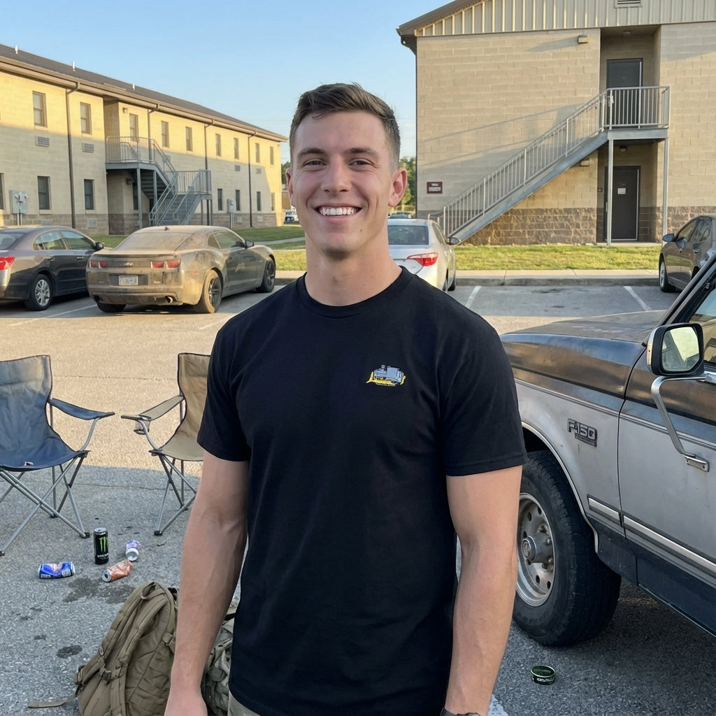 Man in a black t-shirt standing in a military barracks parking lot with cars and chairs around him.