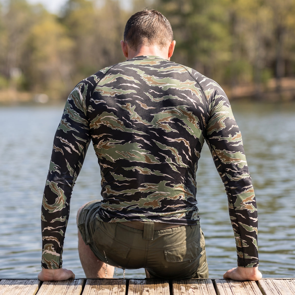 Man wearing a camouflage long-sleeve shirt and shorts, kneeling on a dock by a body of water.