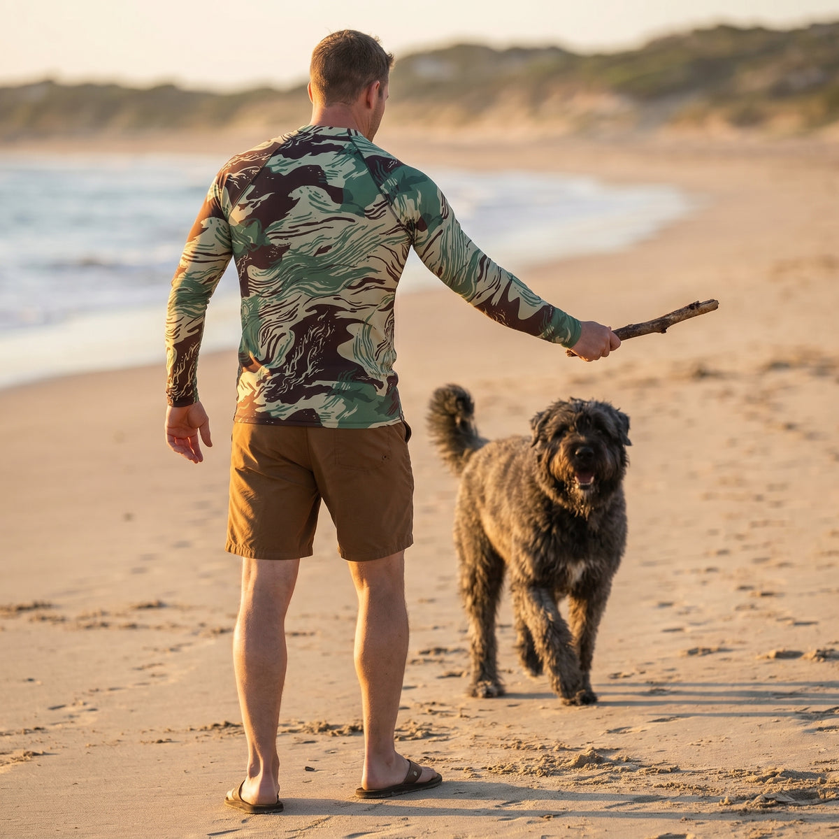 Man in camouflage shirt and shorts standing on a beach with a dog